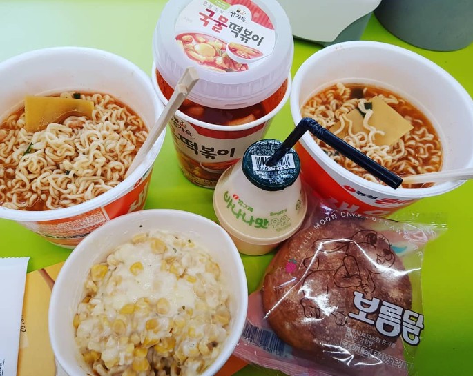Korean convenience store food including triangle kimbap, ramen, lunchbox, and banana milk